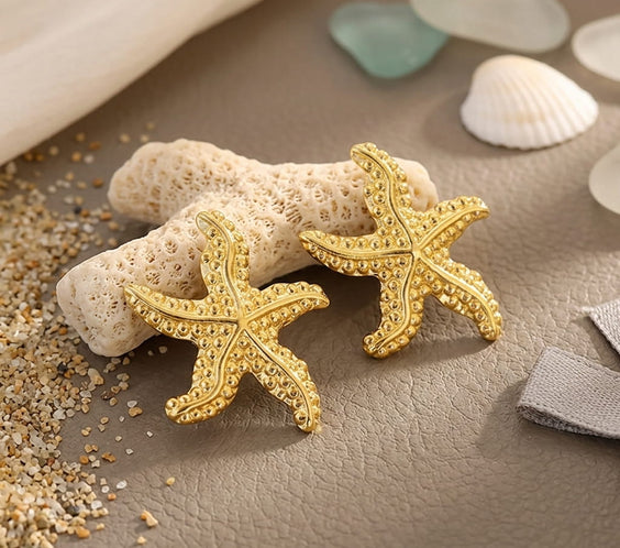 Détail des puces d'oreilles étoiles de mer dorées posées sur un morceau de corail blanc.
