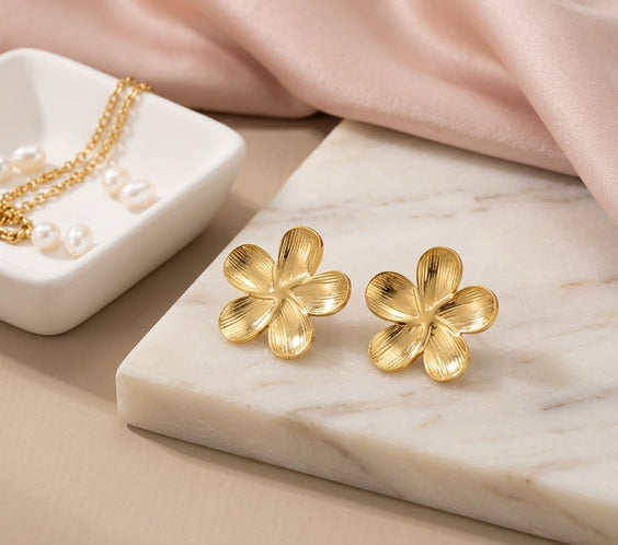 Paire de boucles d'oreilles puces fleur dorées aux pétales striés posées sur une plaque de marbre blanc, avec du tissu satiné rose et un collier de perles.