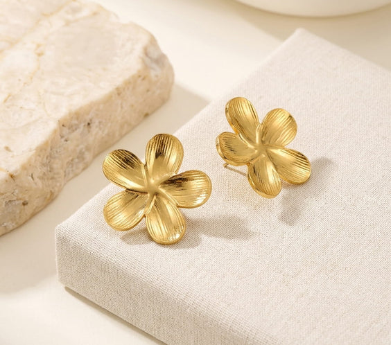 Clous d'oreilles dorés motif fleur texturée présentés sur un présentoir en lin beige à côté d'un bloc en pierre naturelle.
