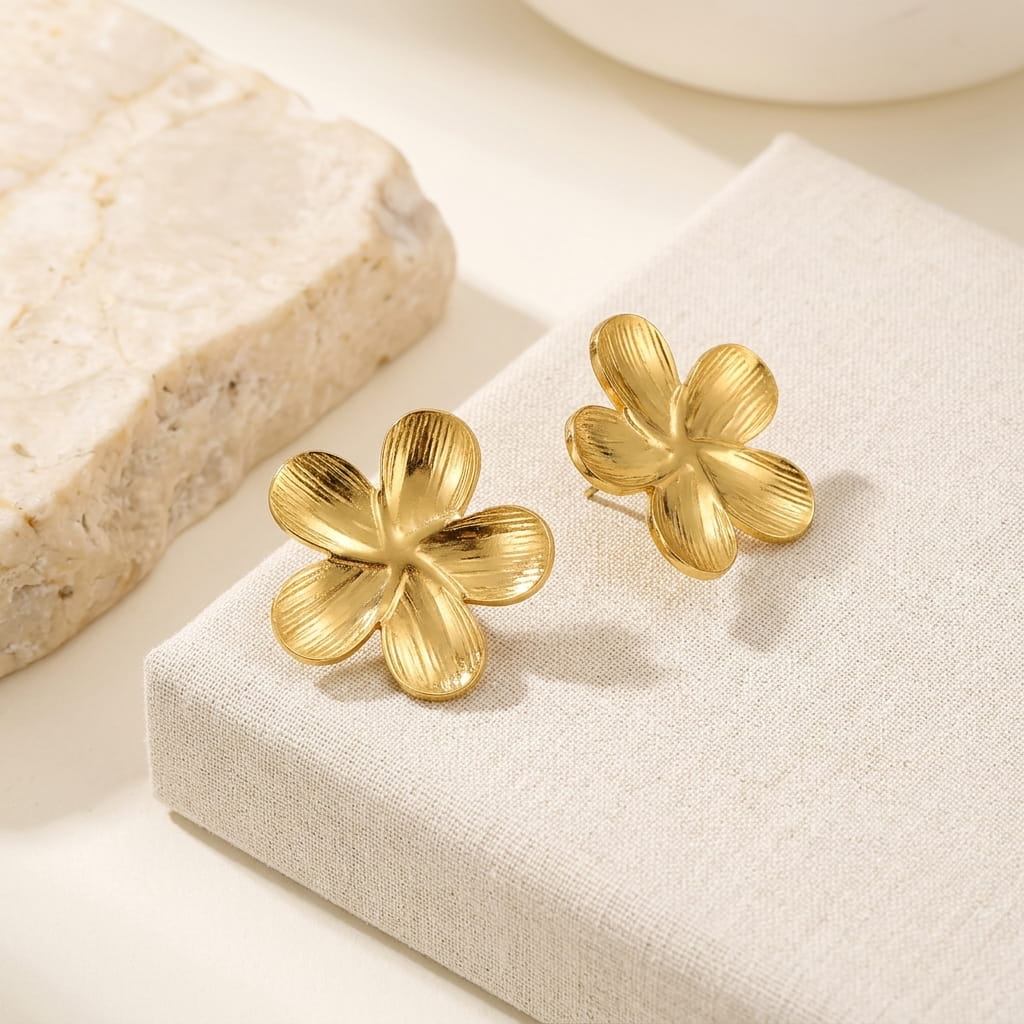 Clous d'oreilles dorés motif fleur texturée présentés sur un présentoir en lin beige à côté d'un bloc en pierre naturelle.