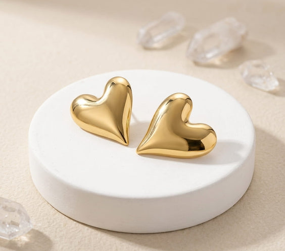 Paire de puces d'oreilles dorées en forme de cœur bombé posées sur un présentoir cylindrique blanc avec des cristaux de quartz.
