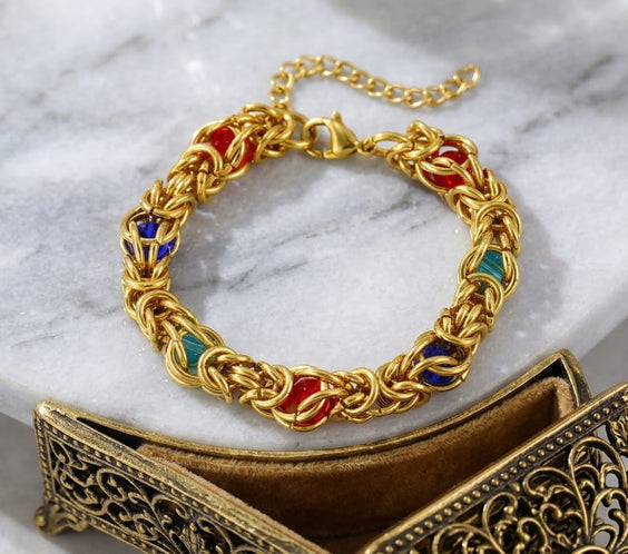 Bracelet maille byzantine doré en acier inoxydable posé sur un plateau en bois avec une fleur blanche.