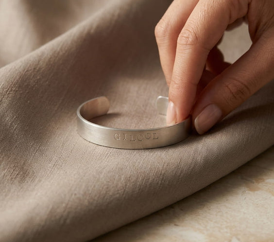 Une main tenant délicatement l'extrémité d'un bracelet jonc argenté brossé gravé d'un mot personnalisé. Le bracelet repose sur un tissu fluide de couleur beige ou taupe, drapé sur une surface en marbre clair.