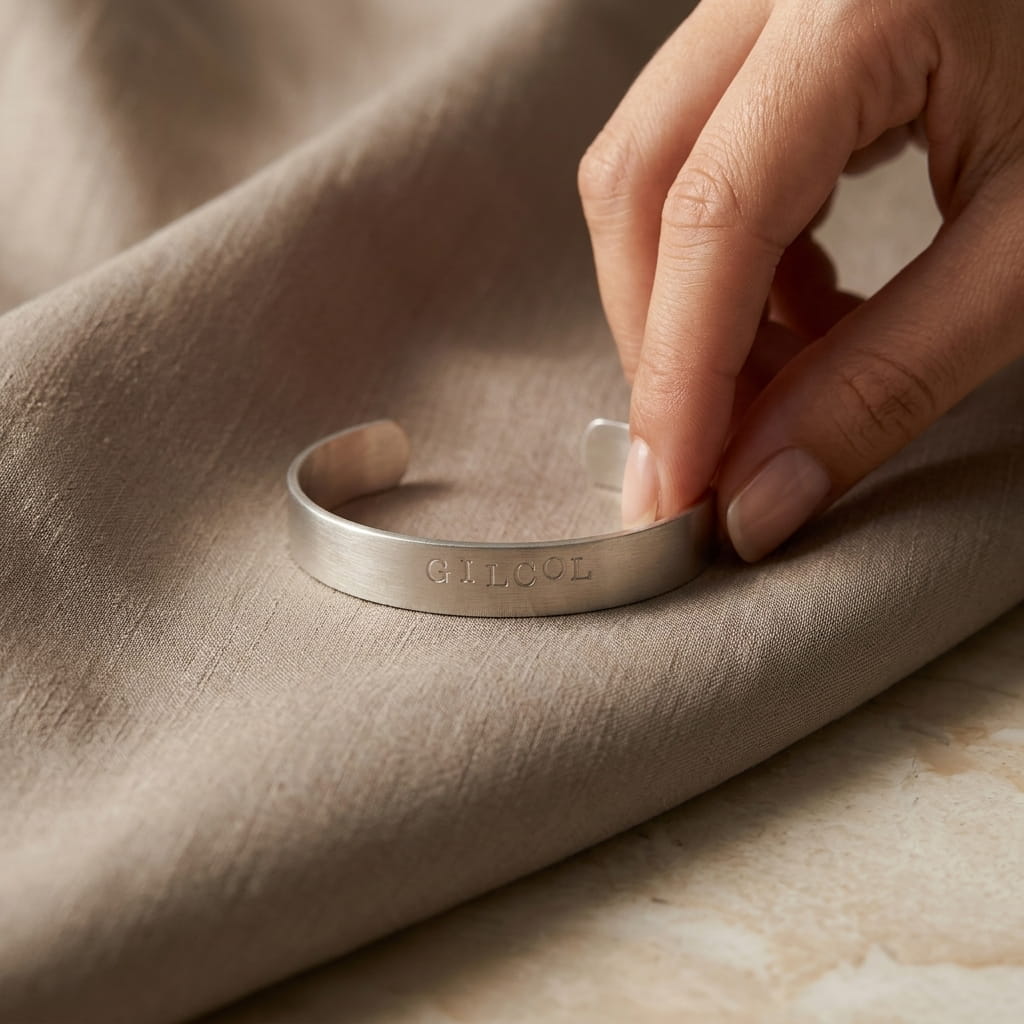 Une main tenant délicatement l'extrémité d'un bracelet jonc argenté brossé gravé d'un mot personnalisé. Le bracelet repose sur un tissu fluide de couleur beige ou taupe, drapé sur une surface en marbre clair.