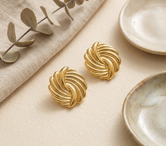 Paire de boucles d'oreilles puces dorées torsadées en acier inoxydable, présentées sur un tissu de lin beige avec des branches d'eucalyptus et des coupelles en céramique artisanales.
