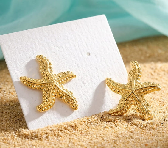 Boucles d'oreilles étoiles de mer dorées présentées sur un support blanc dans un décor de plage.