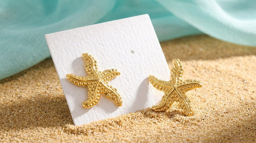 Boucles d'oreilles étoiles de mer dorées présentées sur un support blanc dans un décor de plage.