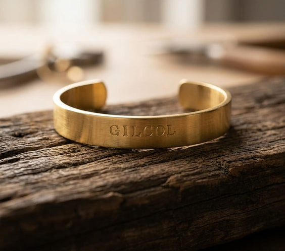Bracelet jonc en métal doré brossé avec la gravure « GILCOL » au centre, posé sur une plaque de marbre blanc aux légères veines grises. Une chaîne en maille dorée serpente en arrière-plan sous une lumière douce.