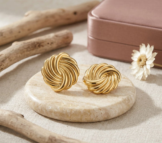 Boucles d'oreilles clous dorées au design sculptural torsadé, posées sur un présentoir rond en marbre avec des branches de bois flotté, une fleur séchée et un écrin cadeau rose poudré en arrière-plan.