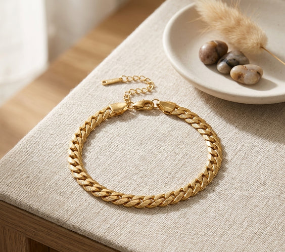Bracelet doré maille serpentine en acier inoxydable posé sur un tissu en lin, accompagné de pierres décoratives et d'un plumeau séché.