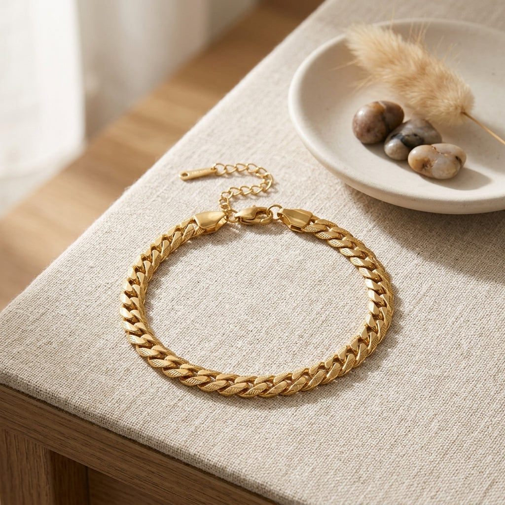 Bracelet doré maille serpentine en acier inoxydable posé sur un tissu en lin, accompagné de pierres décoratives et d'un plumeau séché.