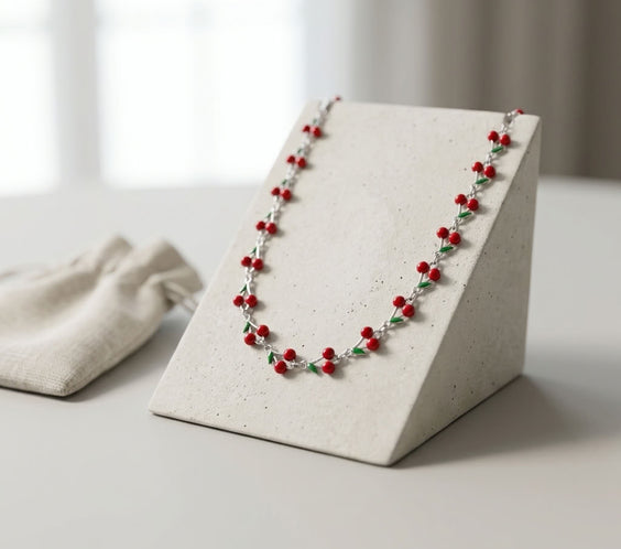 Collier ras de cou argenté avec motifs cerises émaillées rouges et vertes, présenté sur un portant en béton beige à côté d'un pochon en lin.