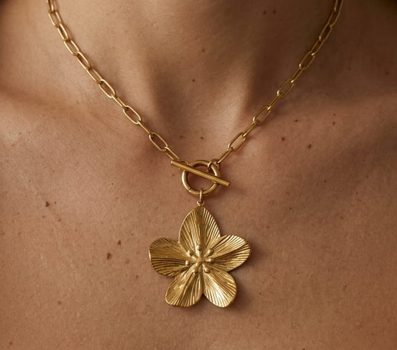 Collier doré avec pendentif fleur texturé sur un fond en lin beige avec fleurs séchées.