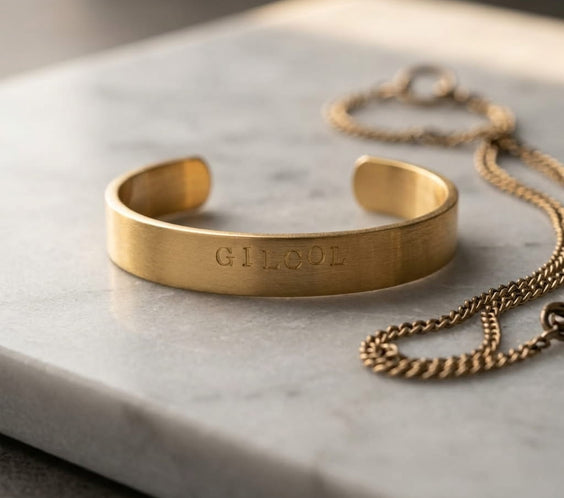 Bracelet jonc rigide en dorure brossée gravé du mot « GILCOL », présenté de face sur un morceau de bois brut et texturé. Arrière-plan flou évoquant l'ambiance chaleureuse d'un atelier d'artisan.