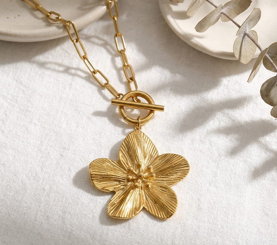 Gros plan du collier maille trombone et pendentif fleur dorée sur surface blanche.