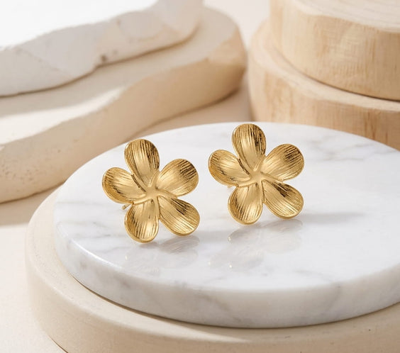 Puces d'oreilles fleur dorées en acier inoxydable posées verticalement sur un socle rond en marbre avec éléments en bois.