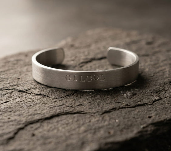 Un bracelet jonc en métal argenté brossé avec l'inscription personnalisée gravée en lettres capitales au centre. Le bracelet est posé horizontalement sur une roche sombre et rugueuse aux textures marquées, sous un éclairage doux et tamisé.